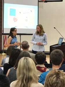 Two high school senior girls stand by a podium and presentation screen and make a presentation.