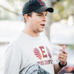 A man with a baseball hat and a tshirt with an E on it talks to high school athletes outside