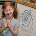 a kindergarten girl stands in front of an easel with a picture she drew